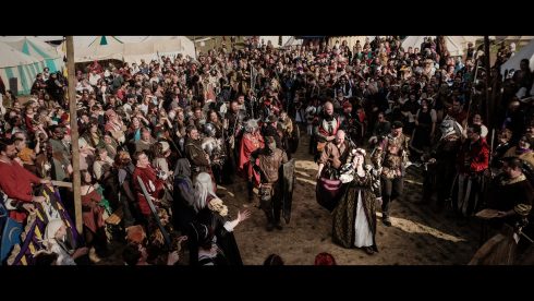 A crowd of people dressed in fantasy costumes line a procession led by a woman in venician-style robes wearing a crown of flowers.