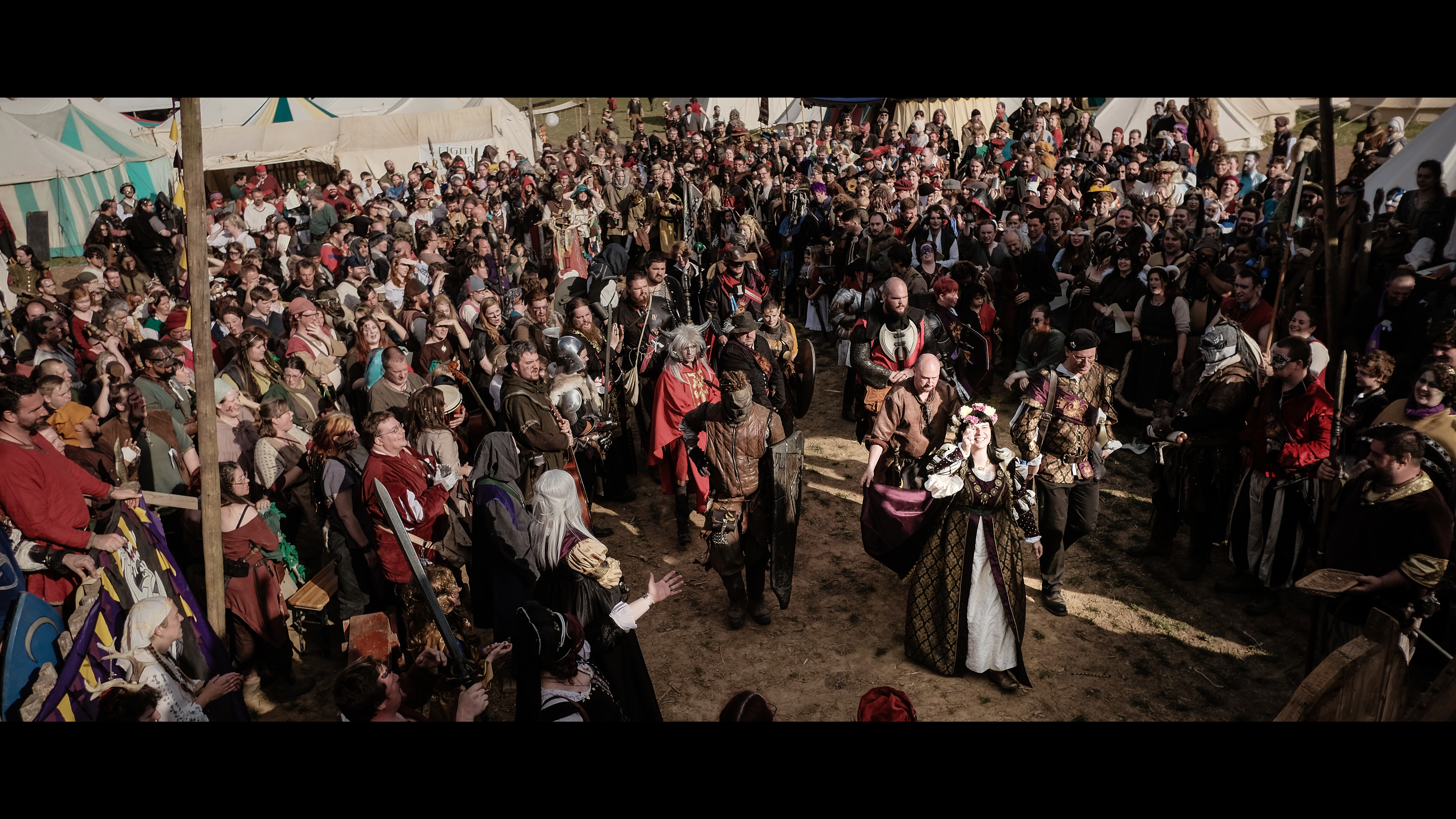 A crowd of people dressed in fantasy costumes line a procession led by a woman in venician-style robes wearing a crown of flowers.